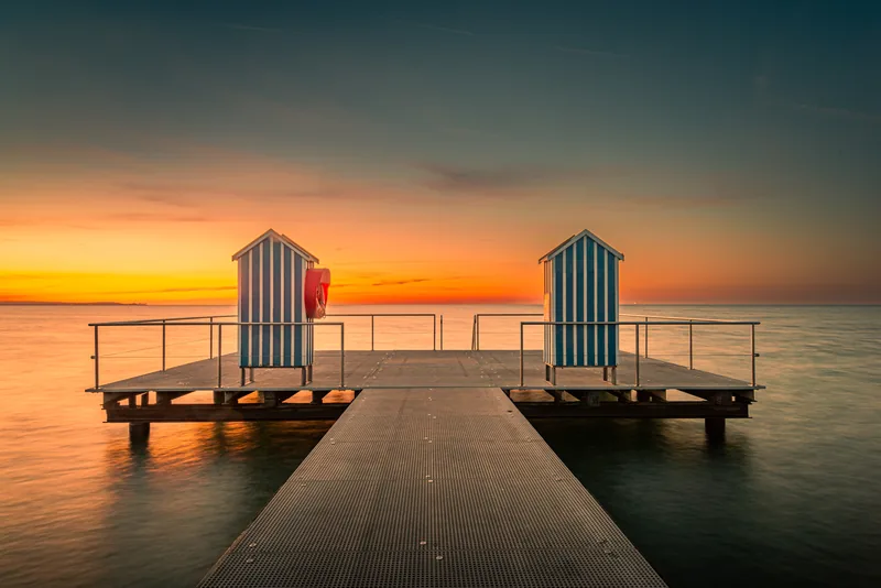 Badesteg mit zwei blau-weiß gestreiften Badehäuschen in der Ostsee, dahinter glüht der Himmel nach Sonnenuntergang