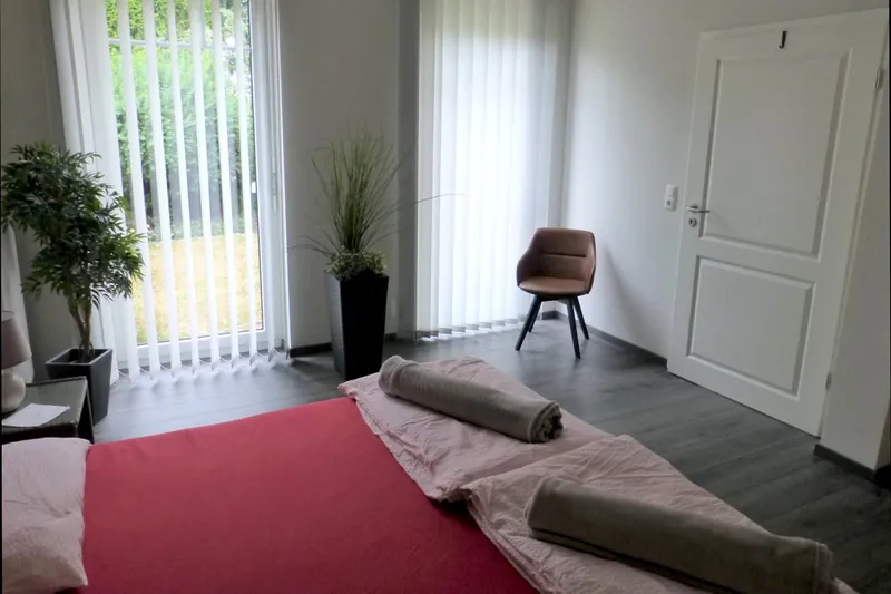 Leseecke mit Sessel neben dem Bett, bodentiefe Fenster mit Lamellenvorhängen zum Garten