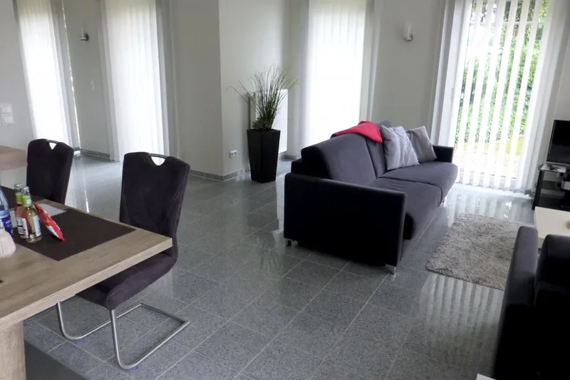 Blick vom Esstisch über den hellen Fliesenboden zum Sofa und den bodentiefen Fenstern