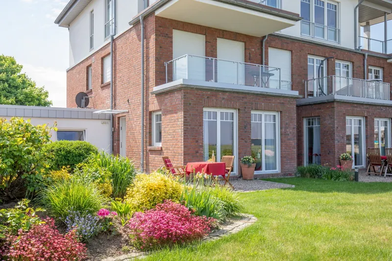 Rotes Klinkerhaus mit Balkonen, davor blühende Beete und Rasen