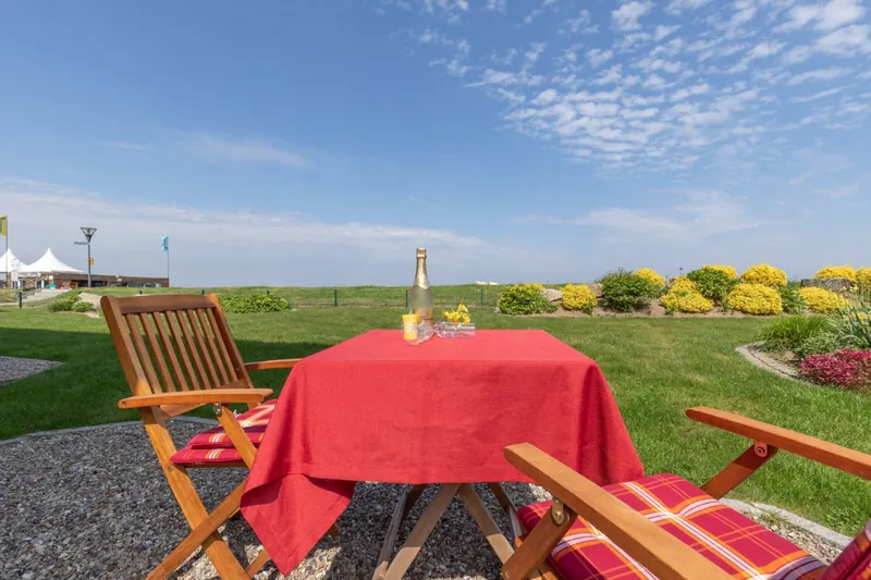 Gartentisch mit roter Decke und zwei Holzstühlen auf der Terrasse, dahinter Rasen, Blumenbeete und Deich
