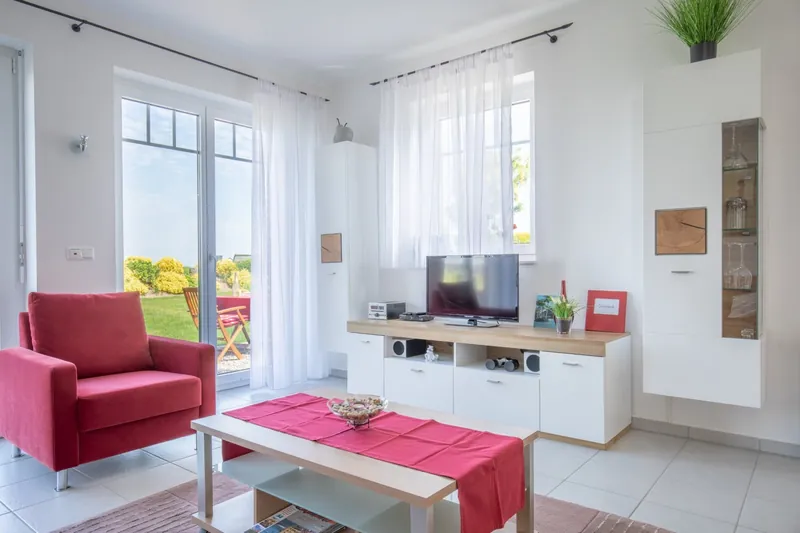 Weißes Lowboard mit Flachbild-Fernseher, roter Sessel und Terrassentür zum Garten