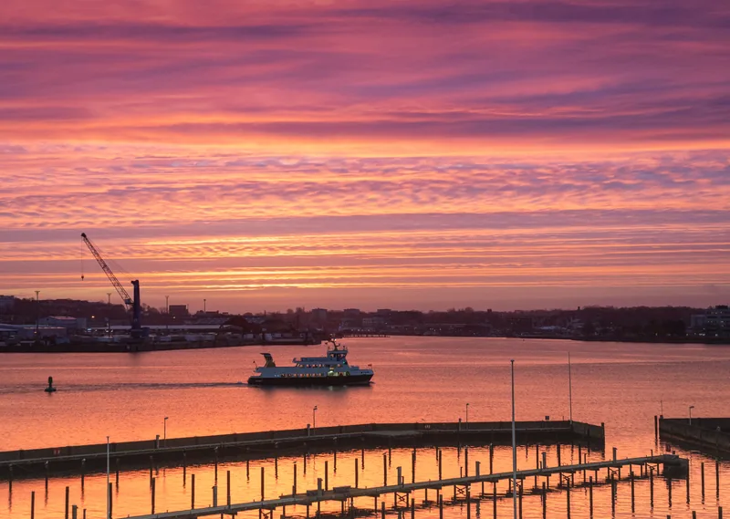 Fördefähre auf der Kieler Förde bei Sonnenaufgang unter rosa-violett leuchtendem Himmel, im Vordergrund Bootsstege