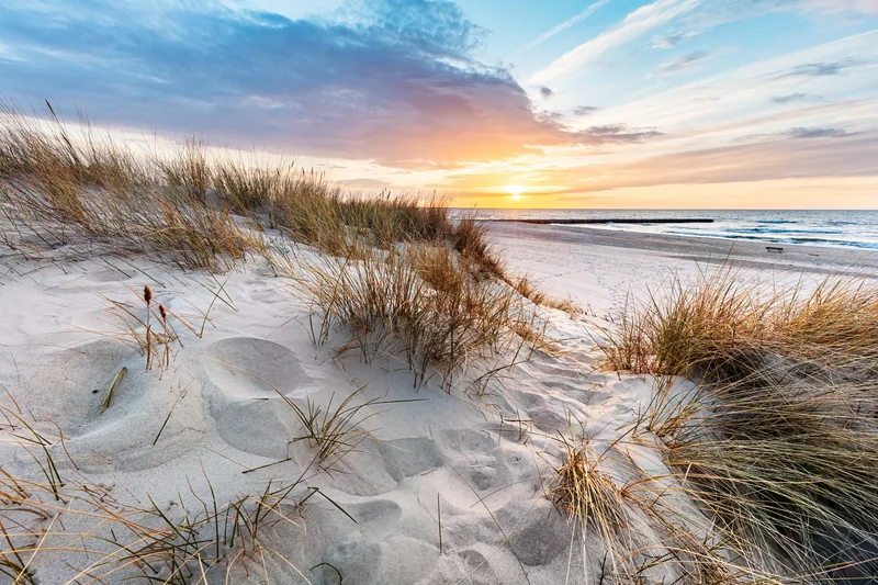 Sanddüne mit Strandhafer und Fußspuren, dahinter Strand und Ostsee bei Sonnenuntergang